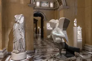 Eine Statue einer weiblichen Figur steht links im Bild, während rechts eine moderne Skulptur eines geflügelten Engels zu sehen ist. Die Umgebung zeigt eine elegante Galerie mit Marmorboden und kunstvollen Wänden, die verschiedene Kunstwerke im Hintergrund präsentiert.