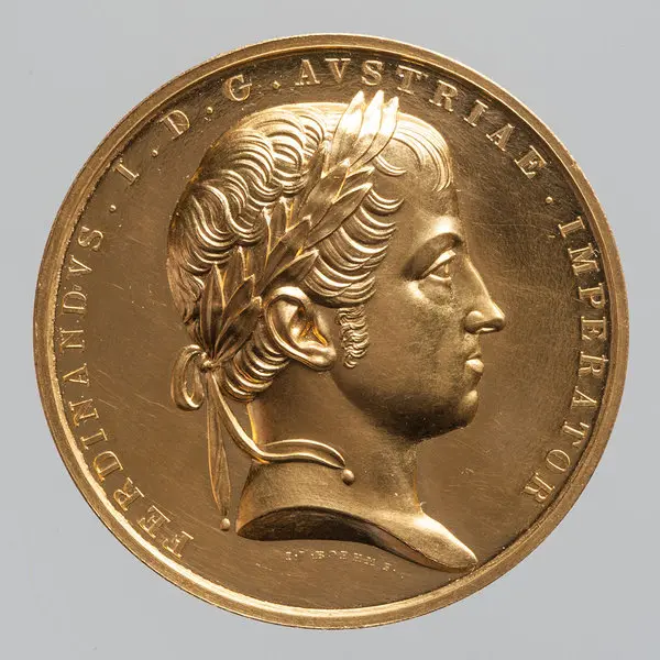 Das Bild zeigt eine goldene Medaille mit dem Profil von Ferdinand I. von Österreich. Er trägt einen Lorbeerkranz und hat einen seriösen Gesichtsausdruck. Um das Bild sind Schriftzüge mit seinem Namen und Titel angebracht.