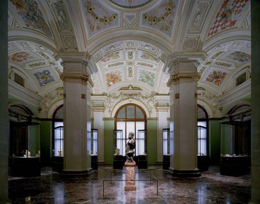 Eine große, kunstvoll gestaltete Galerie mit hohen Decken und bunten Fresken. In der Mitte steht eine Statue auf einem Sockel, umgeben von Vitrinen, die weitere Kunstwerke zeigen. Sanftes Licht fällt durch die Fenster und betont die architektonischen Details der Umgebung.