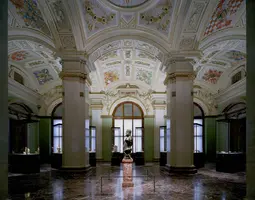 Eine große, kunstvoll gestaltete Galerie mit hohen Decken und bunten Fresken. In der Mitte steht eine Statue auf einem Sockel, umgeben von Vitrinen, die weitere Kunstwerke zeigen. Sanftes Licht fällt durch die Fenster und betont die architektonischen Details der Umgebung.