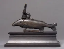 Eine bronzene Skulptur eines fischähnlichen Wesens mit einem aufrechten Horn auf dem Kopf. Der Fisch hat eine angedeutete Rückenflosse und eine geschwungene Körperform. Er steht auf einem rechteckigen Sockel aus Metall.