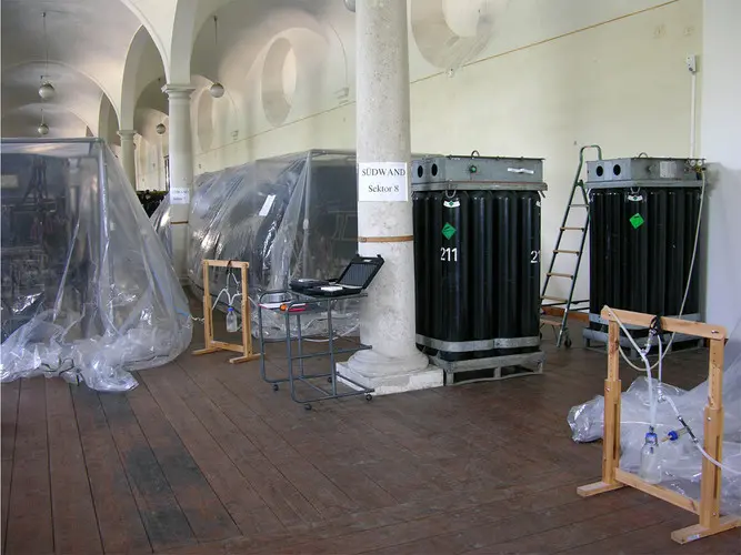 Im Raum sind mehrere große, schwarze Behälter zu sehen, die teilweise mit Plastikfolie abgedeckt sind. Neben einem Behälter steht ein Tisch mit einem Laptop. Es gibt auch eine Holzleiter und einige Holzrahmen im Raum, auf einem Holzfußboden.