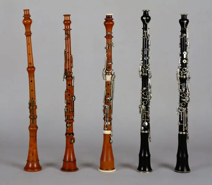 Fünf verschiedene Klarinetten stehen in einer Reihe. Die Instrumente bestehen aus Holz und besitzen unterschiedliche Farben und Verzierungen. Die erste Klarinette ist hellbraun, gefolgt von einer etwas dunkleren, und die letzten drei sind in verschiedenen Schwarz- und Brauntönen gehalten.
