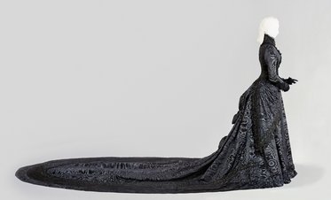 Eine lange, schwarze Robe aus aufwendigem Stoff, die sich bis zum Boden erstreckt. Die Figur trägt die Robe mit einem eleganten Schnitt und einem trächtigen Zug, während das Design durch filigrane Muster verziert ist. Die Silhouette hat blondes, glattes Haar.