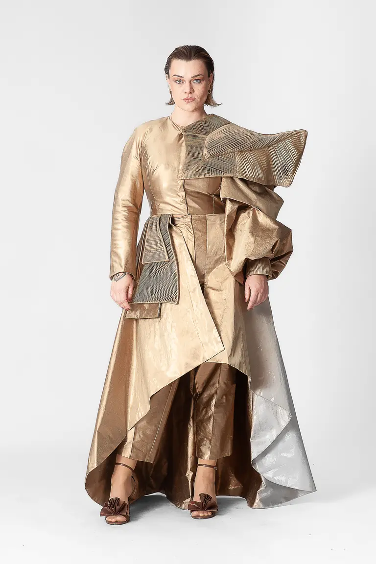 A model stands confidently in a striking golden outfit featuring structured layers and asymmetrical designs. The garment combines shiny fabric with textured elements, creating a bold fashion statement. The model has short hair and wears sandals, posing against a plain white background.