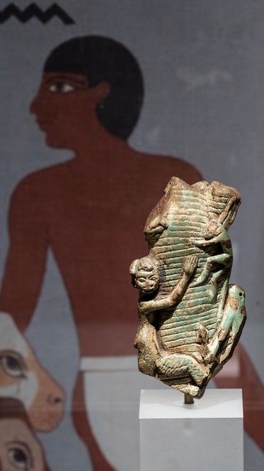 Eine antike ägyptische Skulptur zeigt eine Figur, die einen Menschen darstellt, der an einem Objekt lehnt. Im Hintergrund ist eine Wandmalerei zu sehen, die eine stilisierte menschliche Figur darstellt. Die Skulptur hat eine grüne Patina und steht auf einem weißen Sockel.