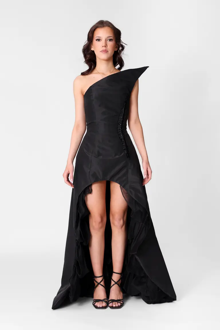 A model stands in a striking black evening gown featuring a one-shoulder design and a high slit. The dress has a dramatic layered hem and is paired with elegant strappy black heels. The background is neutral, emphasizing the gown's unique silhouette.