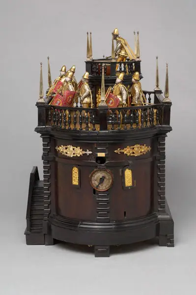 Eine kunstvolle Uhr mit einer runden, dunklen Holzkonstruktion. Auf dem Dach sind prächtig verzierte Figuren von Wachmännern mit Schildern und Helmen angeordnet. Die Uhr hat goldene Verzierungen und ein Zifferblatt auf der Frontseite. Eine Treppe führt zur Uhr.