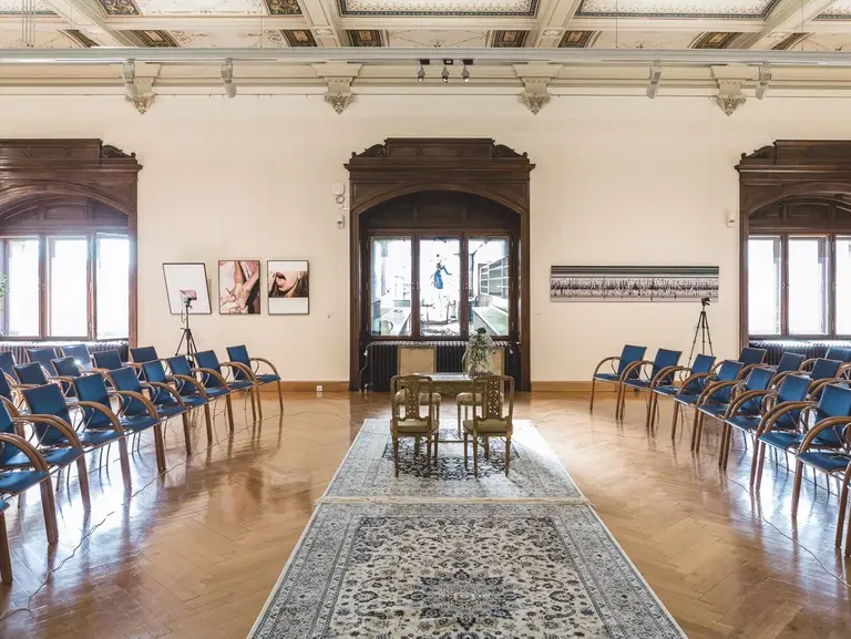 A look into the Bassano Saal of the Kunsthistorisches Museum, that features rows of blue chairs arranged around a decorative rug, with two elegant chairs in the center. The walls showcase framed artworks, and large windows allow natural light to fill the space, enhancing the inviting atmosphere.