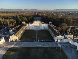 Luftaufnahme eines beeindruckenden historischen Gebäudes mit einer großen, offenen Grünfläche im Vordergrund. Umgeben von Bäumen und Feldern, erstrecken sich die Alpen im Hintergrund, während die Architektur im klassischen Stil harmonisch in die Landschaft eingefügt ist.