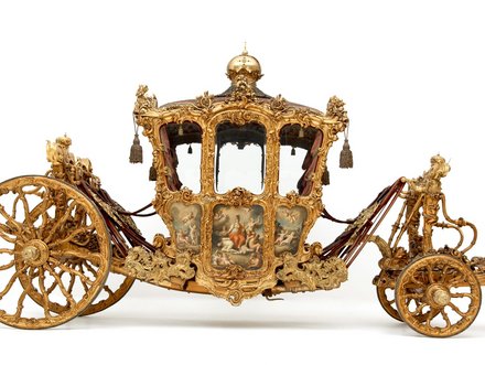 Eine aufwendig gestaltete, goldene Kutsche mit zahlreichen Verzierungen und kunstvollen Details. Sie hat zwei große, dekorierte Räder und eine geschlossene Kabine mit Fenstern, die ein elegantes und prunkvolles Erscheinungsbild vermittelt.