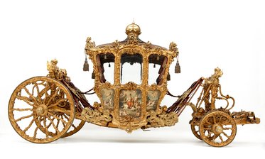 Eine aufwendig gestaltete, goldene Kutsche mit zahlreichen Verzierungen und kunstvollen Details. Sie hat zwei große, dekorierte Räder und eine geschlossene Kabine mit Fenstern, die ein elegantes und prunkvolles Erscheinungsbild vermittelt.