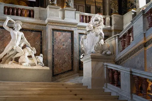 Eine beeindruckende Treppe im KHM mit einer Statue eines Kriegers, der einen Löwen besiegt. Rechts steht eine majestätische Löwenstatue, die ein Wappen hält. Die Wände sind reich verziert mit Marmor und kunstvollen Details. Die Atmosphäre strahlt historische Pracht aus.