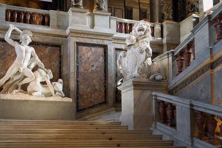 Eine beeindruckende Treppe im KHM mit einer Statue eines Kriegers, der einen Löwen besiegt. Rechts steht eine majestätische Löwenstatue, die ein Wappen hält. Die Wände sind reich verziert mit Marmor und kunstvollen Details. Die Atmosphäre strahlt historische Pracht aus.