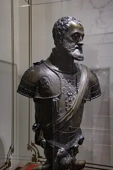 Eine bronzene Büste eines historischen Kriegers, gekleidet in schwerer Rüstung mit detaillierten Verzierungen. Der Krieger trägt einen Bart und hat eine ernsthafte Miene, während er auf die rechte Seite blickt. Die Büste steht in einer gläsernen Vitrine und ist Teil einer Ausstellung.