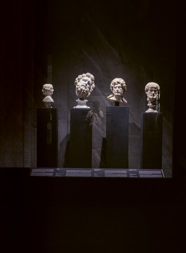 Vier antike Büsten sind in einem lichtgeschützten Ausstellungsraum auf schwarzen Sockeln präsentiert. Die Werke zeigen unterschiedliche Gesichter mit verschiedenen Frisuren und Ausdrücken, beleuchtet von sanftem Licht.