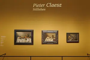 Das Bild zeigt eine Ausstellung mit drei Stillleben-Gemälden von Pieter Claesz auf einer gelben Wand. Der Titel „Pieter Claesz Stillleben“ ist prominent oben sichtbar. Die Gemälde präsentieren verschiedene Szenen mit Obst, Geschirr und anderen Alltagsgegenständen.