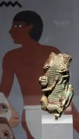 Eine bronzezeitliche Figur, die eine menschliche Gestalt zeigt, die eine andere Figur umarmt, steht auf einem weißen Podest. Im Hintergrund sind Wandmalereien mit einem antiken ägyptischen Motiv zu sehen.