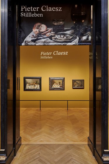 Das Bild zeigt den Eingang zu einer Ausstellung über Pieter Claesz mit einem Banner, das seinen Namen und das Thema "Stillleben" zeigt. Im Hintergrund sind mehrere Gemälde zu sehen, die auf einem goldfarbenen Wandhintergrund hängen. Der Boden ist aus Holz und hat ein Fischgrätenmuster.