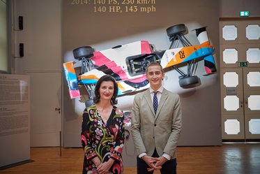 Zwei Personen stehen lächelnd vor einem ausgestellten Rennwagen. Der Wagen ist farbenfroh gestaltet und wird an einer Wand projiziert. Im Hintergrund sind Informationen über den Wagen zu sehen, einschließlich seiner Geschwindigkeit und Leistung.