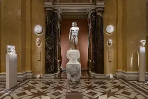 Das Bild zeigt einen kunstvoll gestalteten Museumsraum mit einer zentralen Statue eines männlichen Aktes, umgeben von verschiedenen Skulpturen und Wandreliefs. Der Raum hat elegante Säulen und einen marmorierten Boden, was eine anspruchsvolle Atmosphäre erzeugt.
