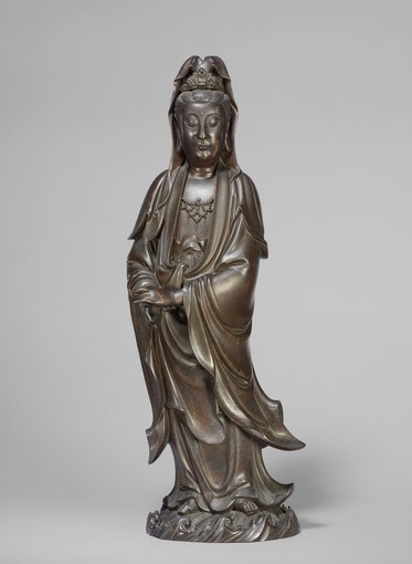 Die Skulptur zeigt eine stehende Figur einer Göttin, die in elegante Gewänder gehüllt ist. Ihr Gesicht strahlt Ruhe und Weisheit aus, während sie eine Hand sanft vor ihrem Körper hält. Die Statue ist aus Bronze gefertigt und betont detailreiche Verzierungen.