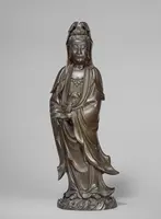 Die Skulptur zeigt eine stehende Figur einer Göttin, die in elegante Gewänder gehüllt ist. Ihr Gesicht strahlt Ruhe und Weisheit aus, während sie eine Hand sanft vor ihrem Körper hält. Die Statue ist aus Bronze gefertigt und betont detailreiche Verzierungen.