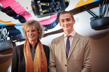 Zwei Personen stehen vor einem ausgestellten Rennwagen. Eine Frau mit lockigem Haar trägt eine schwarze Bluse und einen orangefarbenen Schal. Ein junger Mann in einem hellen Anzug und einer gestreiften Krawatte lächelt. Der Hintergrund zeigt Teile des Rennwagens.