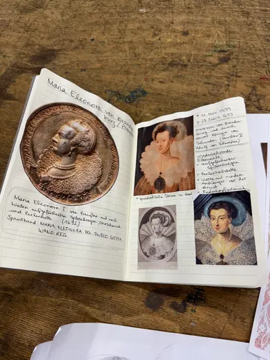 An open notebook displays information about Maria Eleonora, including a medallion image and three portraits. Accompanying text describes her life and significance. The left page features a circular medallion, while the right displays two images of her along with handwritten notes and dates.