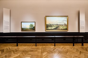 Zwei Gemälde hängen in einer Galerie an einer Wand. Das kleinere Bild zeigt eine Landschaft mit Bäumen, während das größere eine städtische Ansicht darstellt. Eine braune Holzfußbodenfläche ist sichtbar, sowie Informationspulte neben den Bildern.