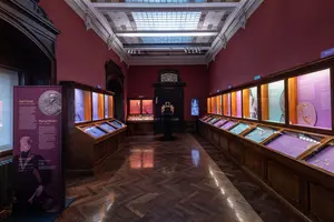Ein Innenraum eines Museums mit rot gestrichenen Wänden. In der Mitte stehen Vitrinen mit verschiedenen Ausstellungsstücken, darunter Münzen und historische Objekte. Auf der linken Seite befindet sich eine Informationswand mit Text und Bildern. Das Licht ist sanft und hebt die Ausstellungsstücke hervor.