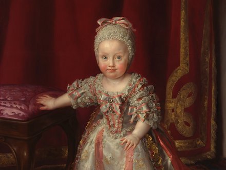 Ein kleines Kind in einem prunkvollen Kleid aus hellen Farben steht neben einer gepolsterten Bank. Das Kind hat einen neugierigen Blick und trägt einen großen Haarreif. Im Hintergrund ist ein roter Vorhang sichtbar, der eine festliche Atmosphäre schafft.