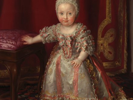 A young child dressed in an ornate gown stands beside an upholstered chair, with a soft expression. The background features rich, deep colors and intricate patterns, enhancing the regal atmosphere of the portrait. The child’s light-colored hair is adorned with a ribbon.
