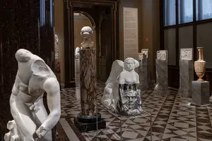 Der Raum zeigt eine Galerie mit verschiedenen Marmorskulpturen. Im Vordergrund stehen mehrere Skulpturen auf Sockeln, darunter eine Engelfigur und ein bärtiger Gesichtsausdruck. Die Wände des Raumes sind mit erklärenden Texten geschmückt, und das Licht beleuchtet die Kunstwerke sanft.