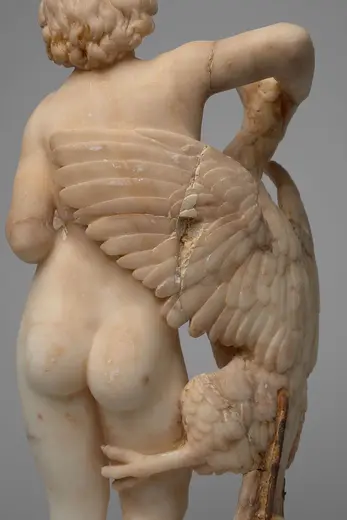 Die Skulptur zeigt von hinten einen nackten Jungen mit lockigem Haar. Er hält einen Vogel mit ausgebreiteten Flügeln in seiner rechten Hand. Der Fokus liegt auf der feinen Detailarbeit des alabasterartigen Materials und der Pose des Jungen, die Anmut und Zartheit vermittelt.