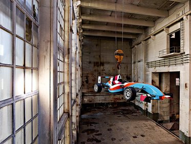 Ein Formelrennwagen in leuchtenden Farben hängt an einem Kran in einer alten, weitläufigen Industriehalle. Die Wände sind rustikal und verfallen, mit großen Fenstern, die Licht in den Raum lassen. Der Boden ist uneben und zeigt Spuren von Wasser oder Rost.