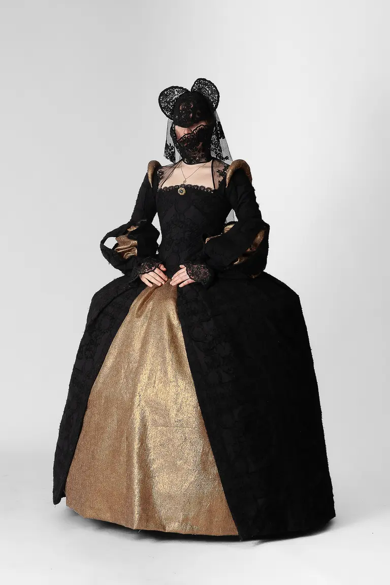 A model wearing a dramatic historical-inspired gown features a fitted black bodice with ornate sleeves and a voluminous gold skirt. A black lace veil and matching headpiece complete the look, evoking a sense of elegance and mystery against a plain white background.
