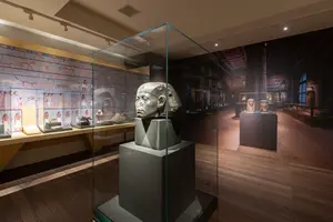 Eine antike Skulptur eines Kopfes in einem gläsernen Ausstellungsstück. Im Hintergrund sind Wandmalereien und weitere Artefakte in einem modernen Museum sichtbar. Die Beleuchtung betont die Details der Skulptur und der Ausstellung.