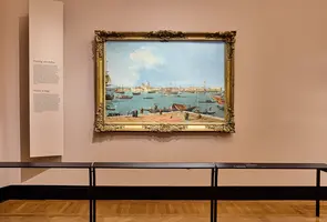 Ein Gemälde in einem goldenen Rahmen zeigt einen Hafen mit Schiffen und Menschen am Ufer. Hinter dem Bild stehen Informationsschilder an der Wand, die den Besuchern Details über das Kunstwerk und den Künstler bieten. Der Boden ist aus Holz und die Wände sind in sanften Farben gehalten.
