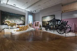 Im Museum ist ein Raum mit historischen Kunstwerken zu sehen. Darunter zwei prunkvolle Wagen, einer goldfarben und einer schwarz mit kunstvollen Verzierungen. An der Wand hängen große Gemälde, die beeindruckende Szenen zeigen. Der Boden ist aus Holz und der Raum ist gut beleuchtet.