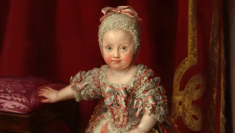 Ein kleines Kind in einem üppigen historischen Kleid mit Rüschen und Schleifen steht neben einem gepolsterten Stuhl. Das Kind hat einen sanften Gesichtsausdruck und einen hellen Teint. Im Hintergrund ist ein rotes Vorhangmotiv sichtbar, das einen eleganten Rahmen bietet.