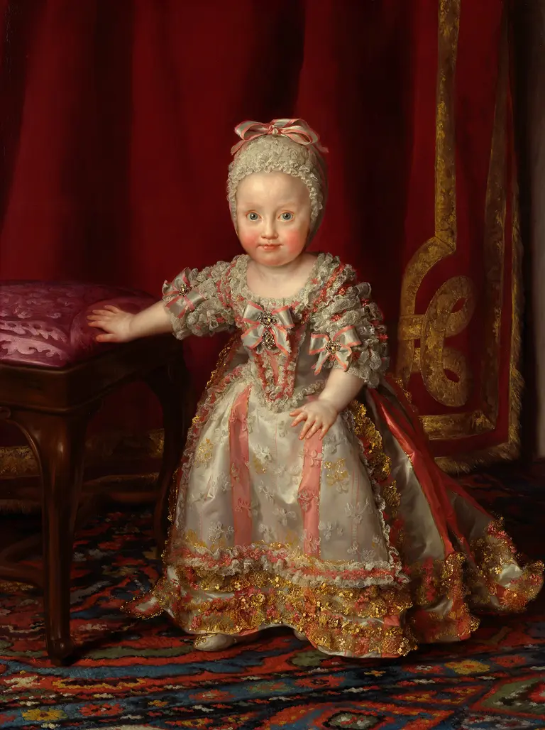 Ein kleines Kind in einem üppigen historischen Kleid mit Rüschen und Schleifen steht neben einem gepolsterten Stuhl. Das Kind hat einen sanften Gesichtsausdruck und einen hellen Teint. Im Hintergrund ist ein rotes Vorhangmotiv sichtbar, das einen eleganten Rahmen bietet.