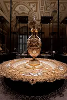 Ein prächtiges goldenes Gefäß steht auf einem kunstvoll verzierten Unterteil. Das Gefäß hat filigrane Details und einen hohen Aufbau mit einer dekorativen Spitze. Es ist von elegantem Design und präsentiert sich in einem stilvollen Raum mit historischer Architektur.
