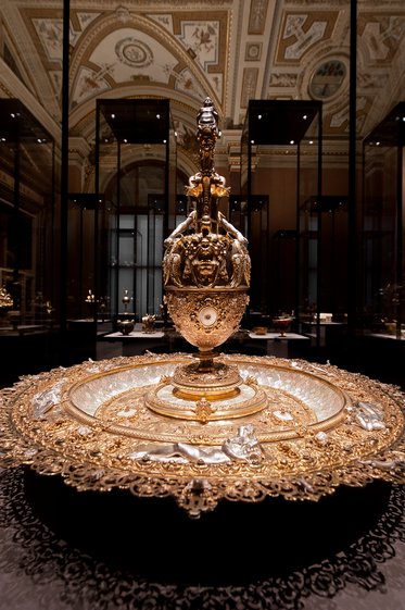 Ein prächtiges goldenes Gefäß steht auf einem kunstvoll verzierten Unterteil. Das Gefäß hat filigrane Details und einen hohen Aufbau mit einer dekorativen Spitze. Es ist von elegantem Design und präsentiert sich in einem stilvollen Raum mit historischer Architektur.