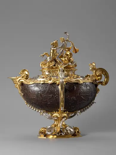 Ein kunstvoll verziertes Gefäß aus Silber und Gold, von dem ein Figurenpaar, das einen Fackelträger darstellt, gekrönt wird. Das Gefäß hat geschwungene Verzierungen und ist auf einem dekorativen Fuß platziert. Die Oberfläche zeigt filigrane Muster.