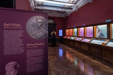 Das Bild zeigt eine Ausstellung mit einem roten Hintergrund und beleuchteten Vitrinen. Auf der linken Seite steht ein großes Plakat mit dem Titel „Kopf und Kragen – Männern machen Mode“. In den Vitrinen sind verschiedene Exponate zur Modegeschichte zu sehen.