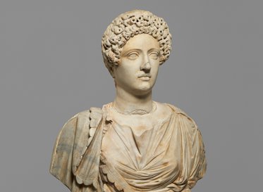 Eine antike Marmorstatue einer Frau mit einer detailliert ausgearbeiteten Frisur und einem Gewand. Sie hat einen neutralen Gesichtsausdruck und ist auf einem grauen Hintergrund platziert.