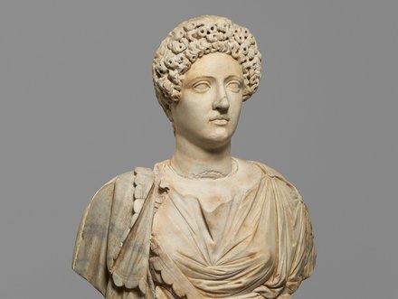 Eine antike Marmorstatue einer Frau mit einer detailliert ausgearbeiteten Frisur und einem Gewand. Sie hat einen neutralen Gesichtsausdruck und ist auf einem grauen Hintergrund platziert.
