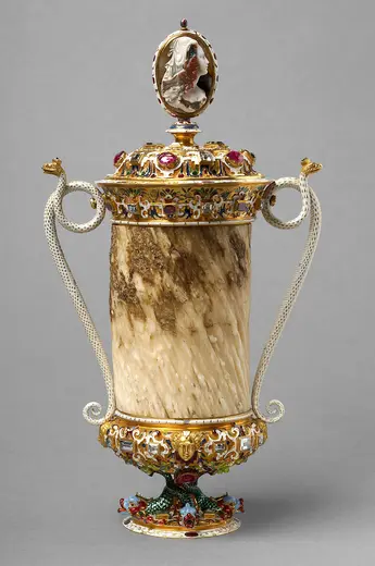 Eine kunstvoll verzierte Vase aus Marmor mit einem oberen Deckel, der eine Büste zeigt. Sie hat goldene Verzierungen und farbige Edelsteine, sowie zwei seitliche Henkel. Die Unterseite ist reich verziert und zeigt eine kreative Gestaltung.