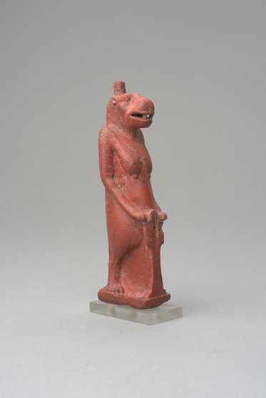 Eine kleine Statue eines menschenähnlichen Körpers mit einem Tierkopf, möglicherweise eines Hundes oder einer anderen Kreatur. Die Statue ist aus rotem Material und steht auf einer transparenten Basis. Der Kopf ist leicht nach vorne geneigt, und die Figur hält ein Objekt in der Hand.